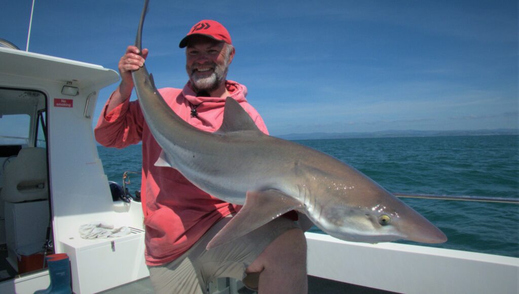 Epic Fishing 2 On Epic Fishing trips in Cardigan Bay, Julian-Lewis-Jones holds up a shark aboard a boat off New Quay, Wales, with the sea calm behind him.